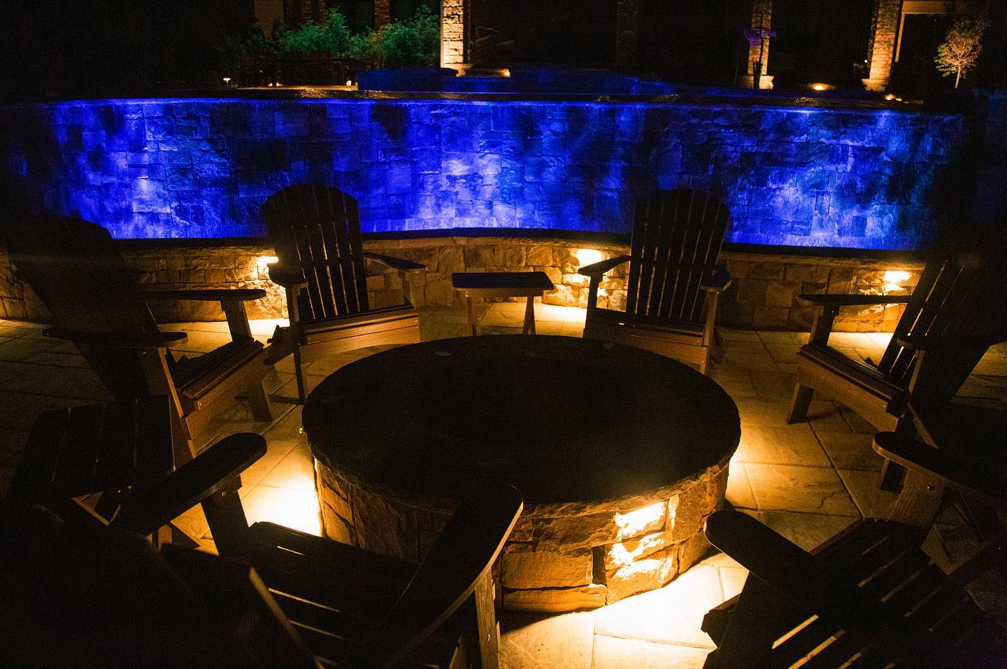 Lit fire pit surrounded by wooden chairs, with blue-lit waterfall backdrop at night.