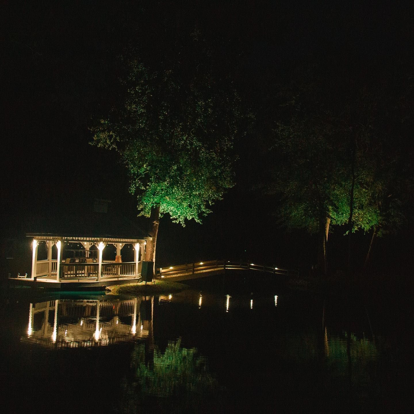 Lit gazebo by a dark pond at night, with glowing tree reflections on the water