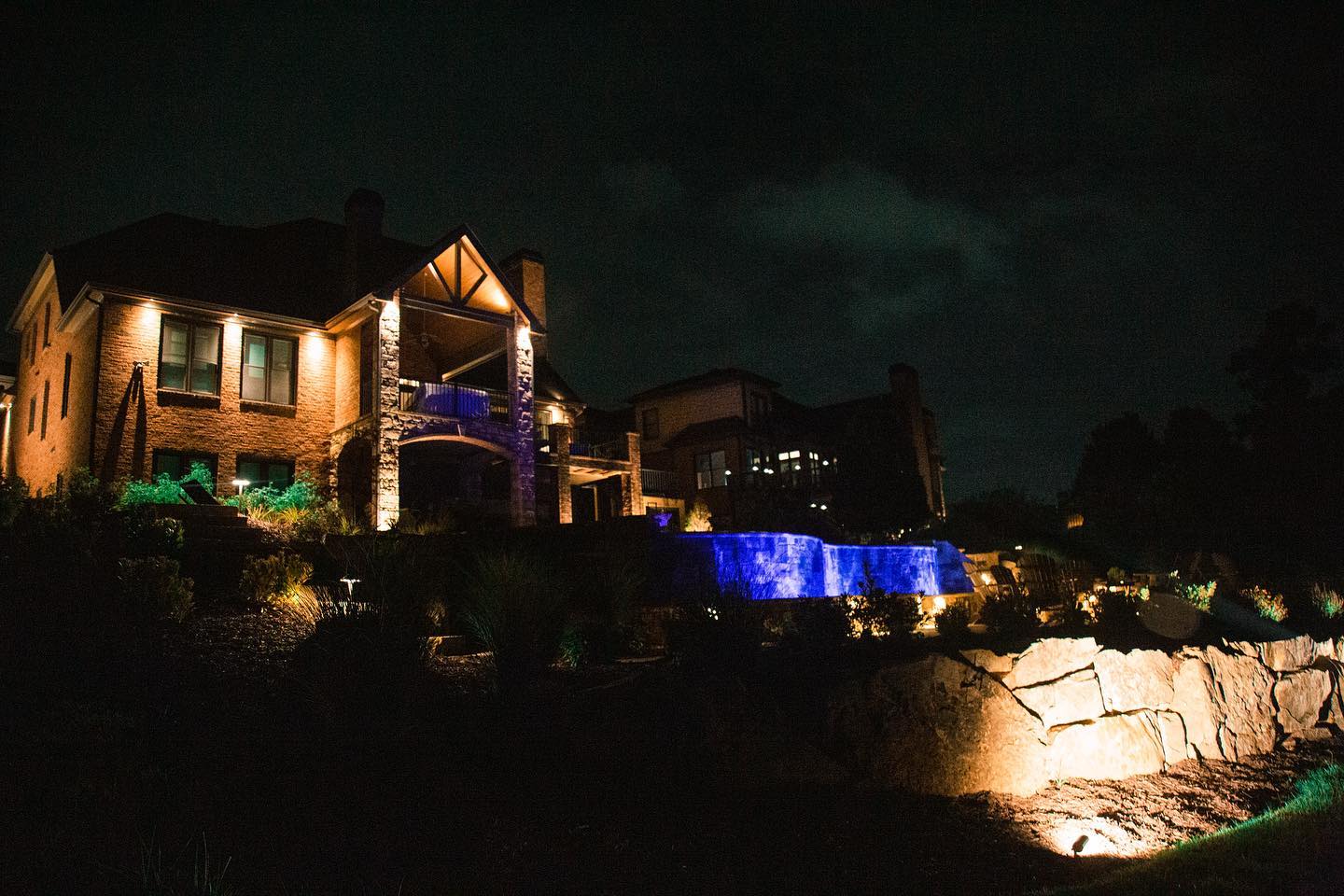 Nighttime view of a lit stone house and blue-lit pool in a landscaped yard