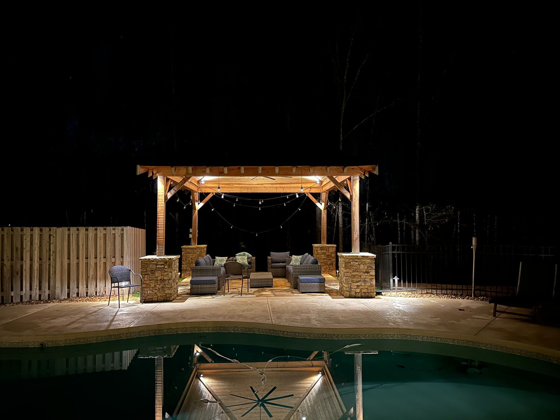 Lit backyard pool at night with a wooden pergola and lounge chairs beside a fence.