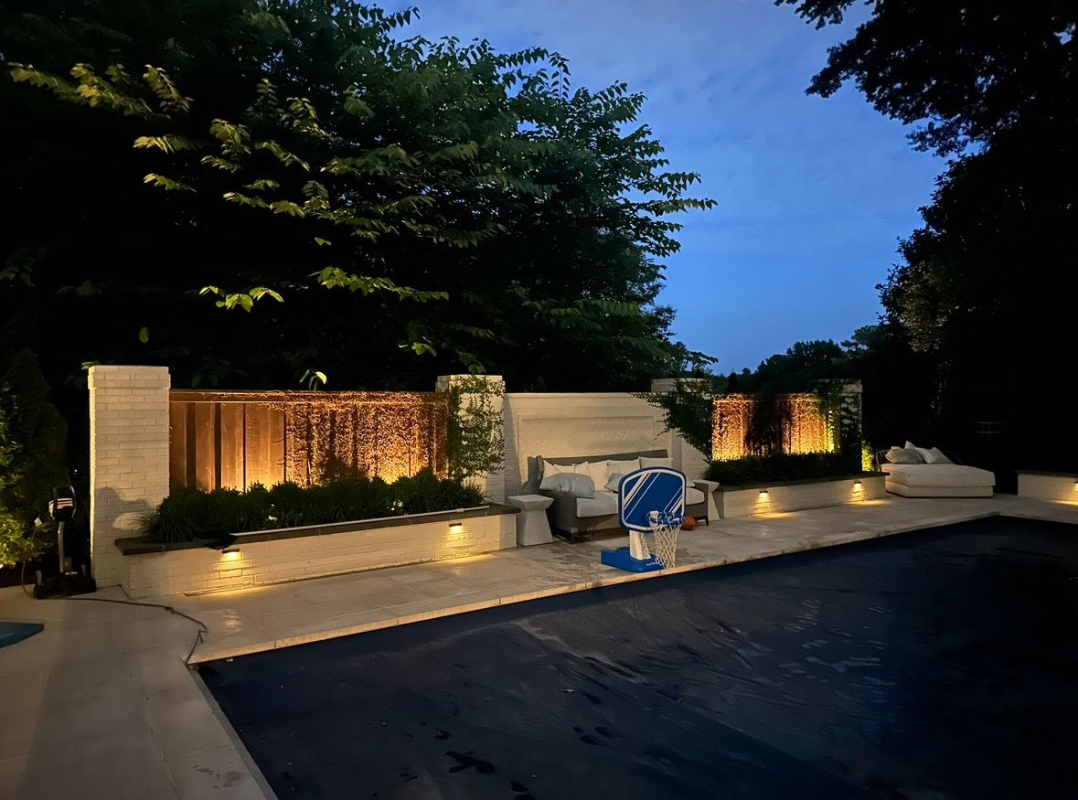 Lit outdoor patio with water feature, low walls, and trees at dusk