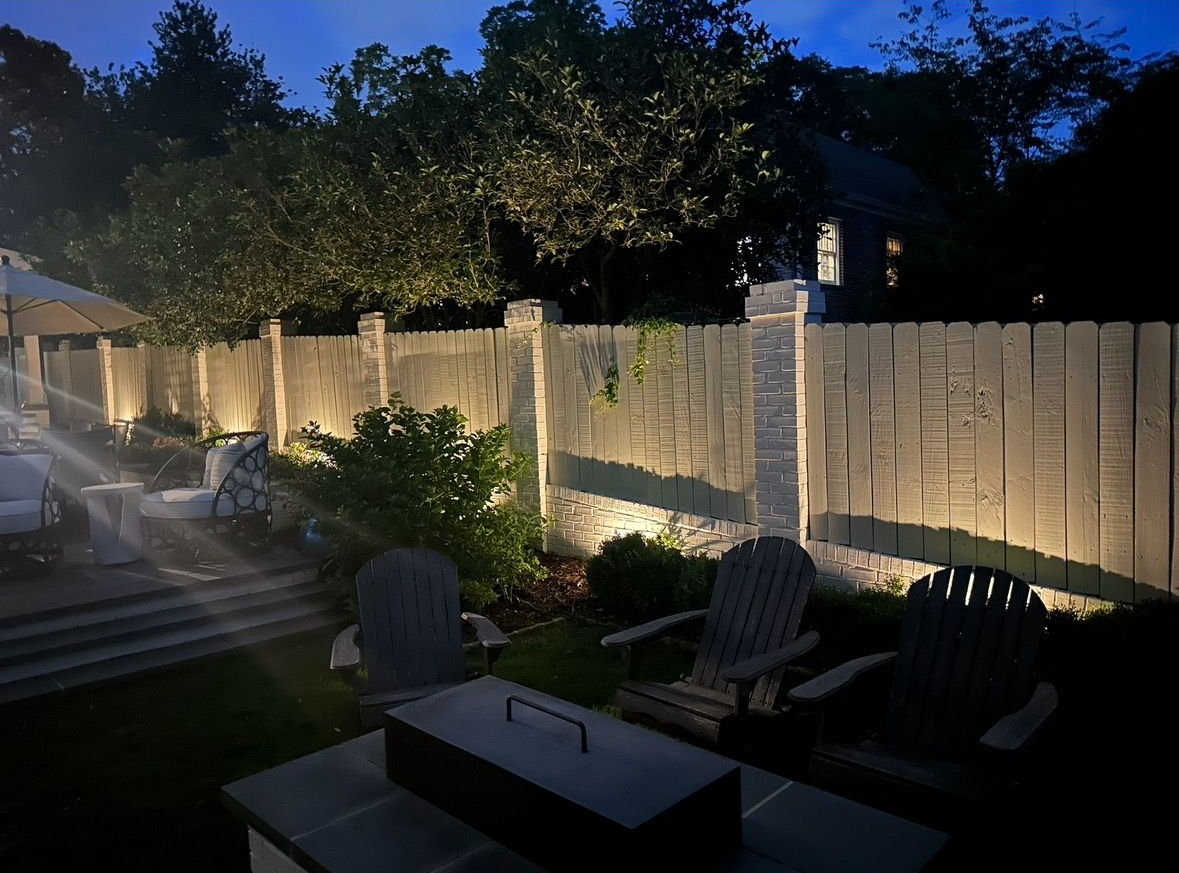 Backyard patio at dusk with string lights, wooden fence, chairs, and a fire pit