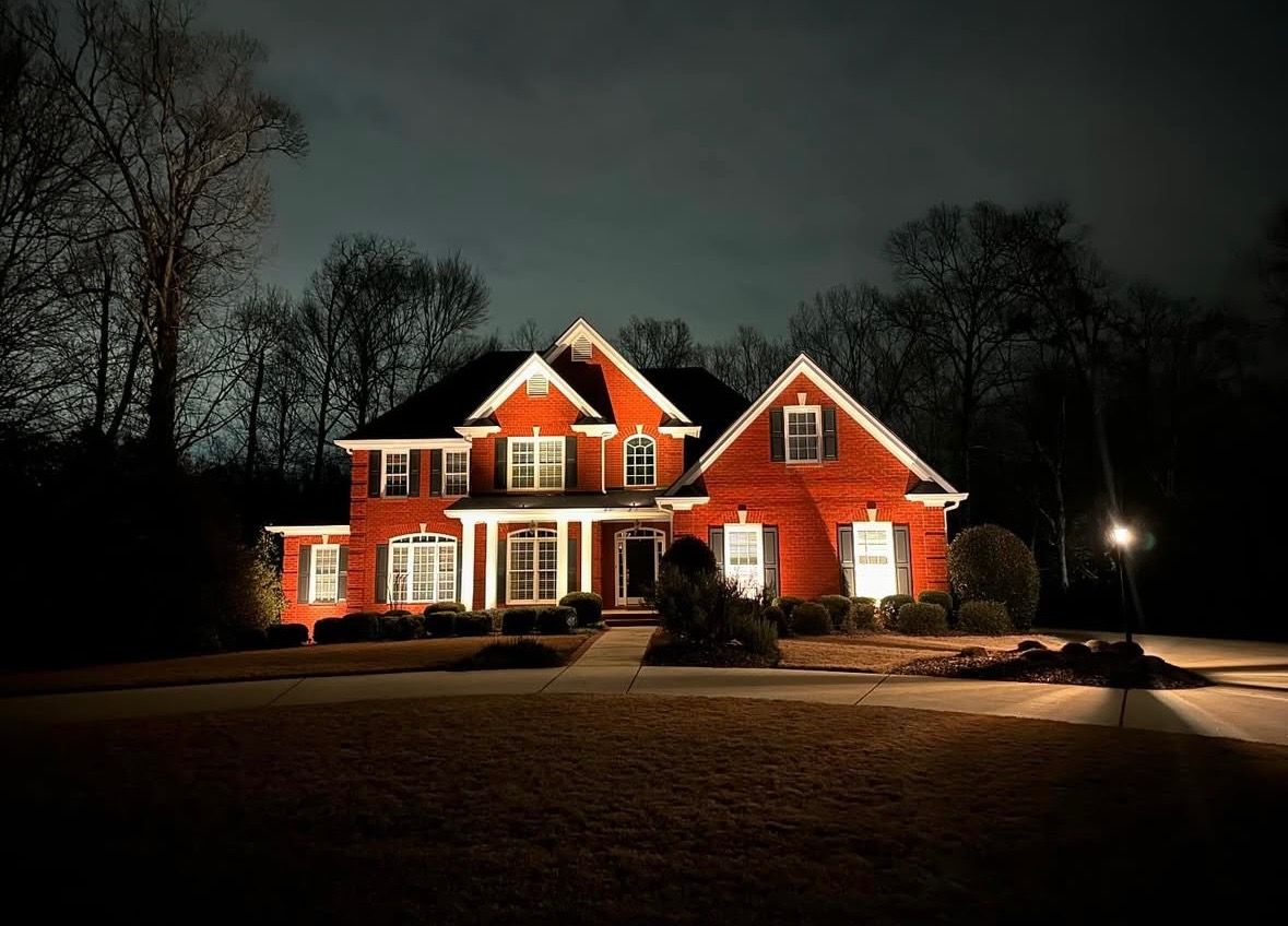 Red brick house lit at night, with dark trees and a driveway under a cloudy sky