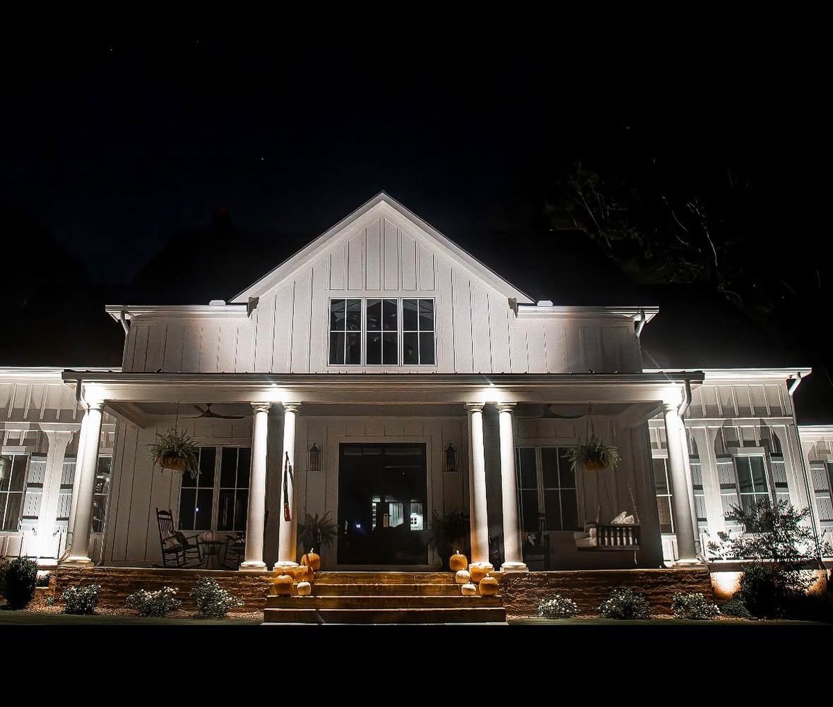 Well-lit white house at night with a peaked roof and front porch, surrounded by landscaping