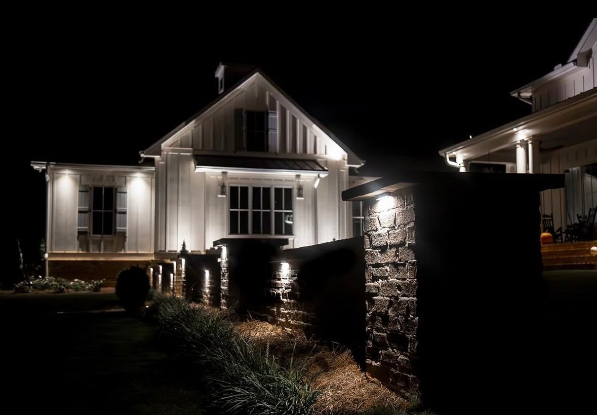 Nighttime view of a lit suburban house with porch lights and a stone pillar in the foreground