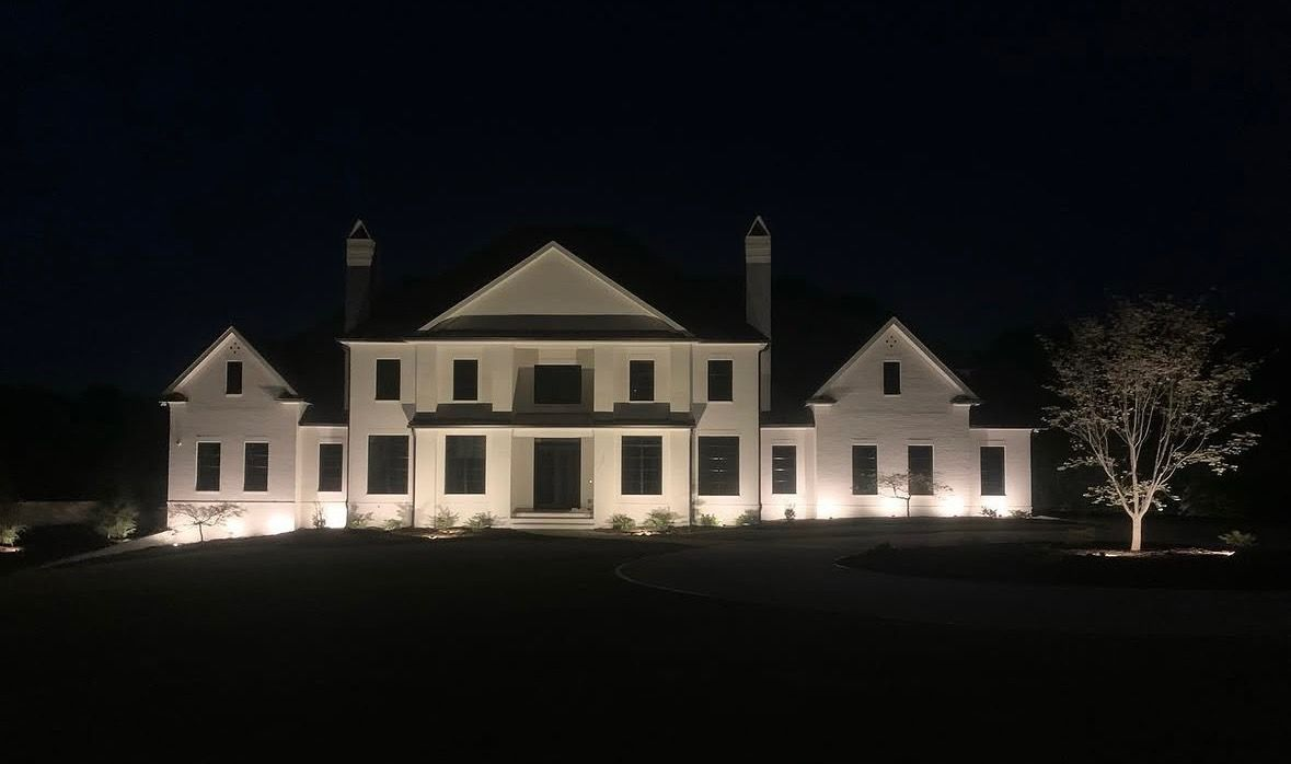 Illuminated white two-story mansion at night with trees on the lawn and a dark sky.
