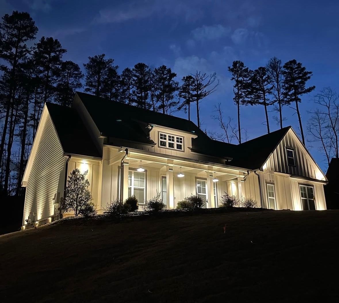 Illuminated house at dusk with white columns and tall pine trees silhouetted against a deep blue sky