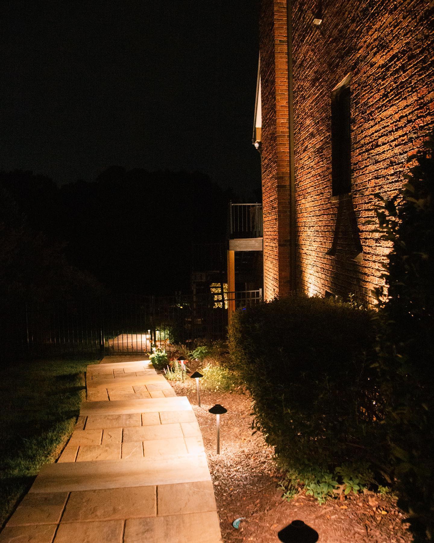 Lit stone walkway beside a brick building at night, with bushes and path lights illuminating the scene