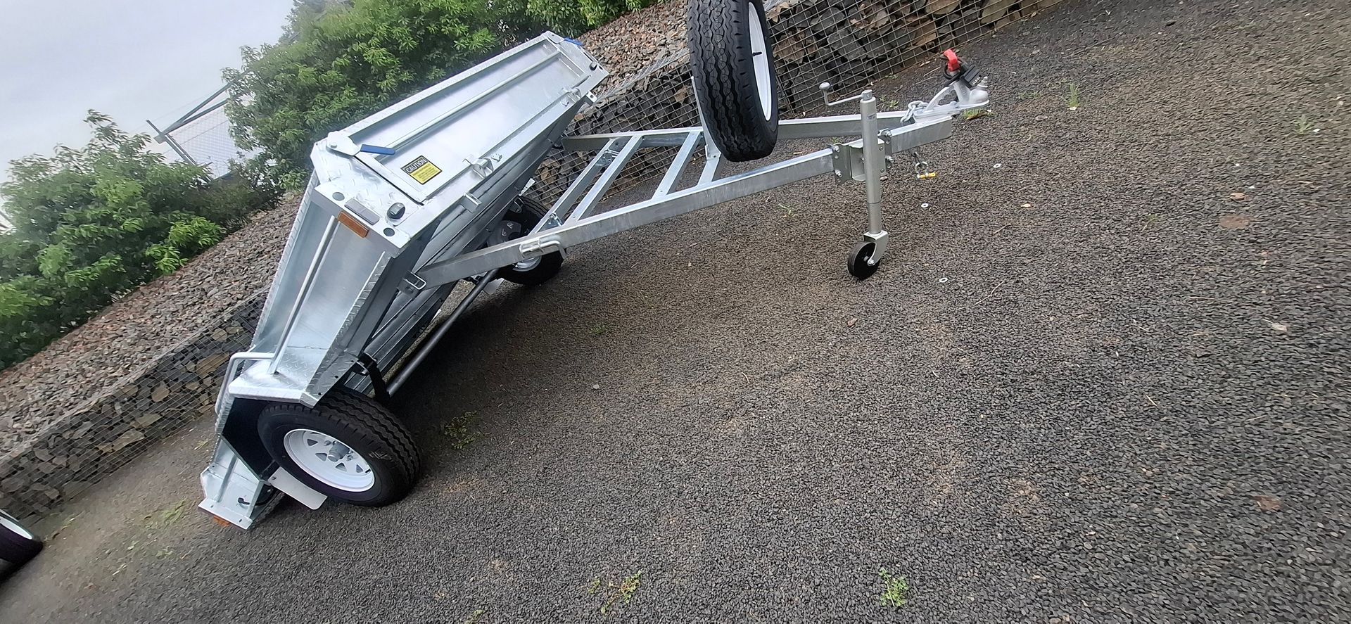 A silver utility trailer with a tilted bed on a gravel driveway — Towform Trailers in Brocklehurst, NSW