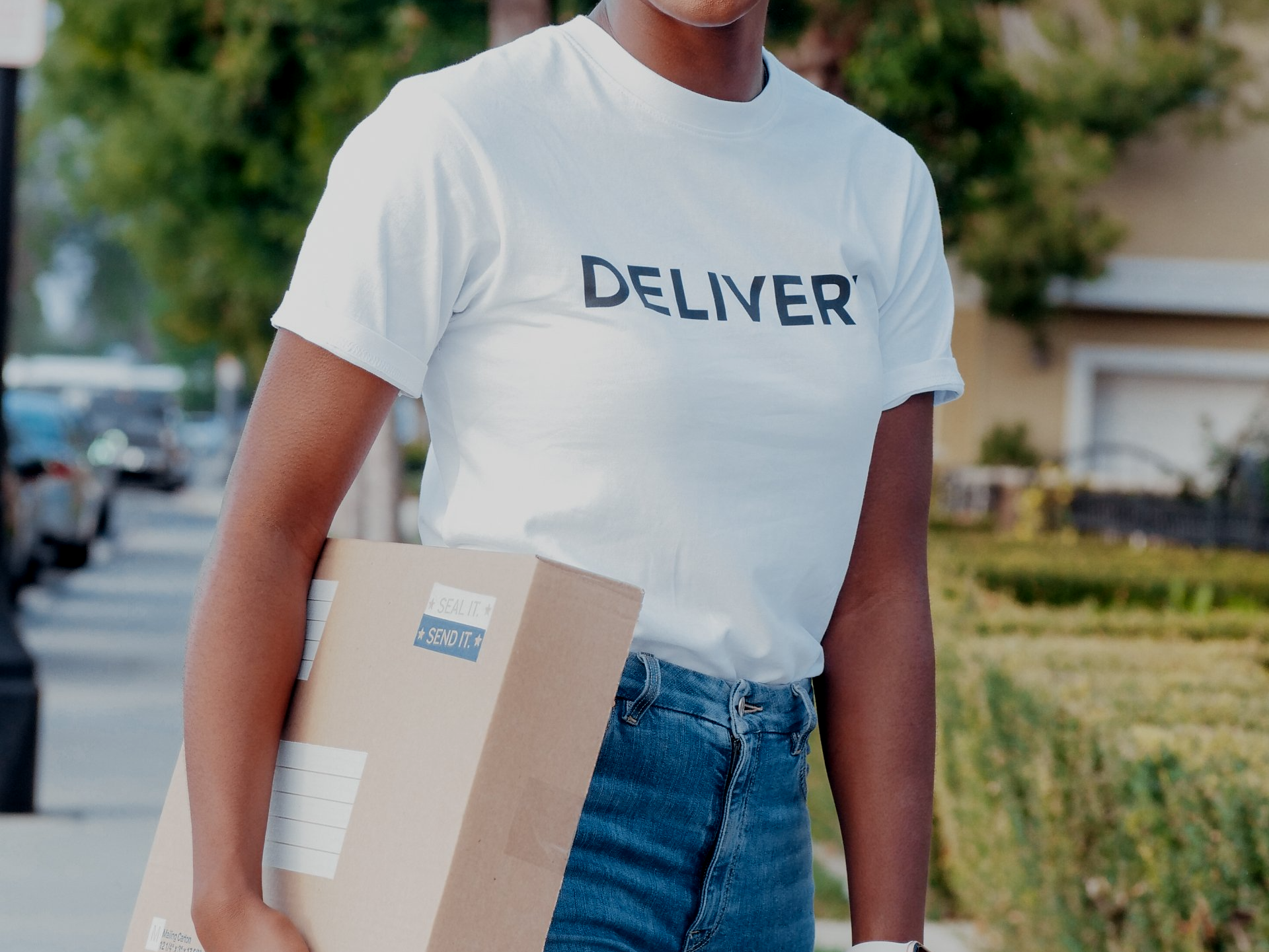 A woman wearing a white t-shirt that says `` deliver '' is holding a cardboard box.