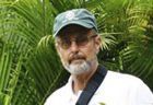 Man in a cap and glasses standing among green tropical plants, wearing a white shirt and black strap