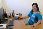 Woman smiling at a desk in an office, wearing a blue top beside a computer and papers