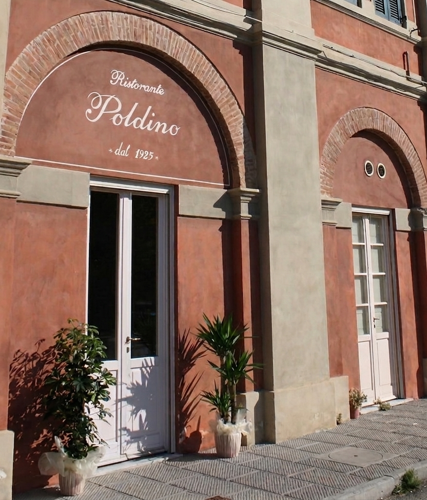 Ingresso del ristorante con portale ad arco, porta bianca aperta e insegne sul marciapiede su una facciata rosa.