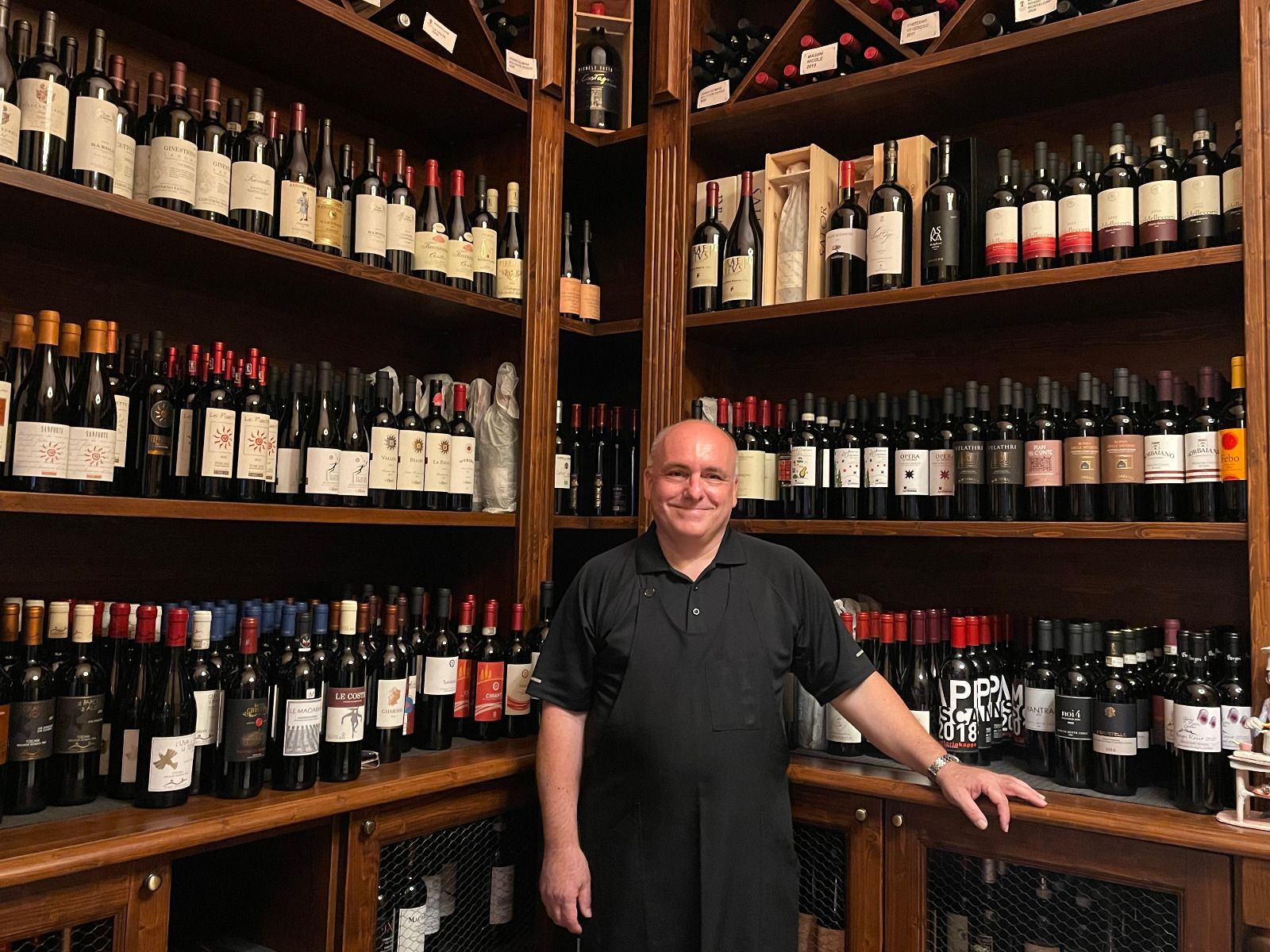 Un uomo in piedi in una cantina, circondato da scaffali pieni di bottiglie di vino.