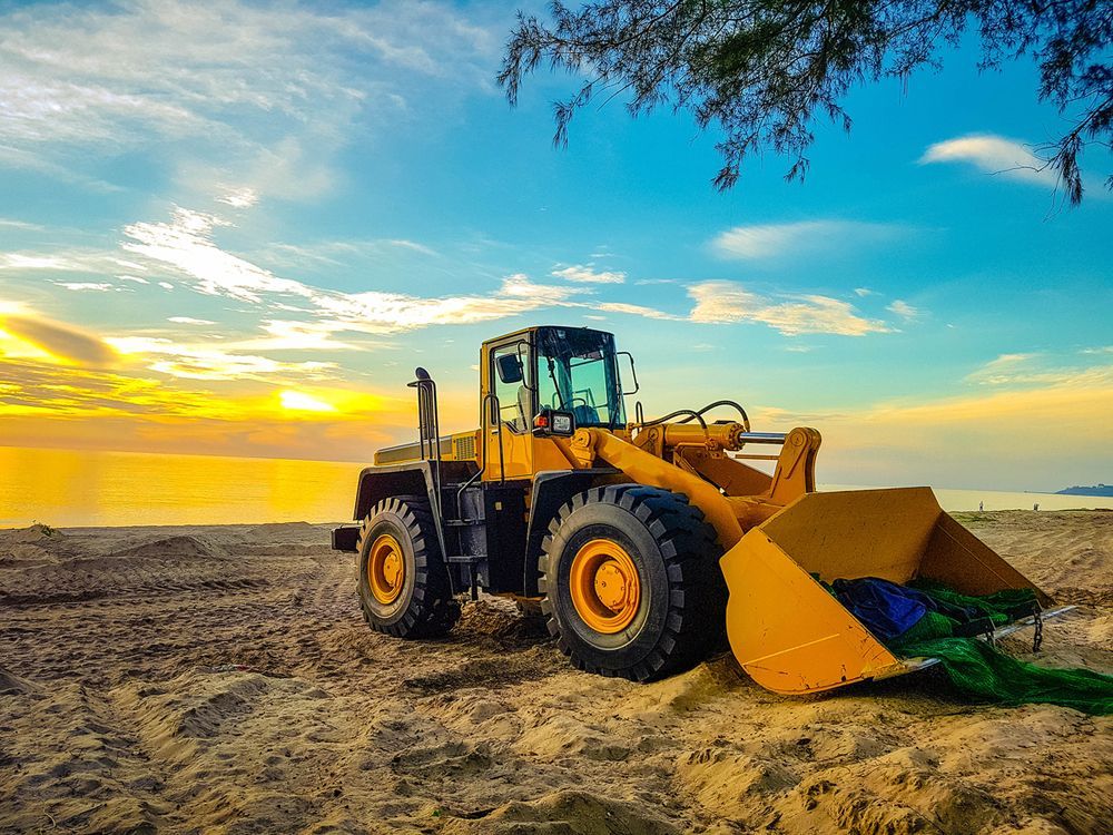 A Yellow Bulldozer Is Parked on The Beach at Sunset — PBC Compliancing in Moranbah, QLD