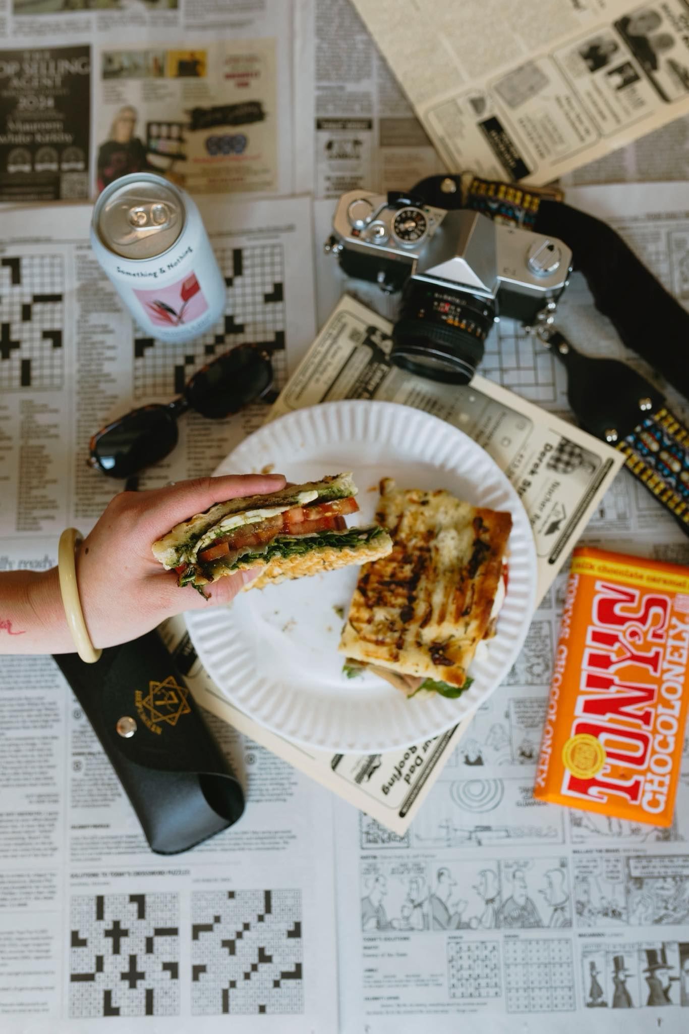 Sandwich on a paper plate, hand holding a half. Newspaper background with a camera, soda, sunglasses, and chocolate bar.