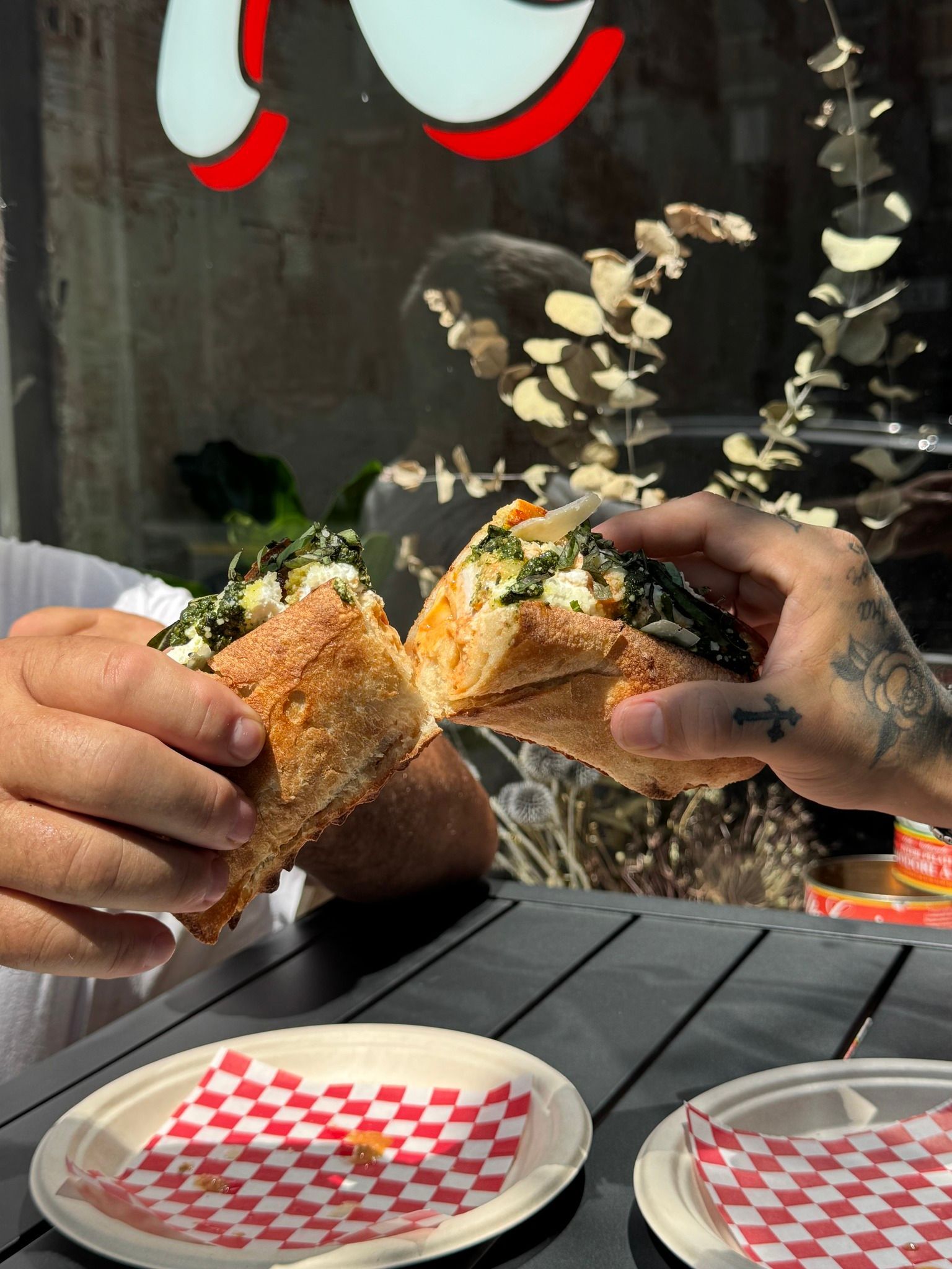 Two people holding halves of a sandwich outdoors at a table, near a window, checkered napkins on plates.