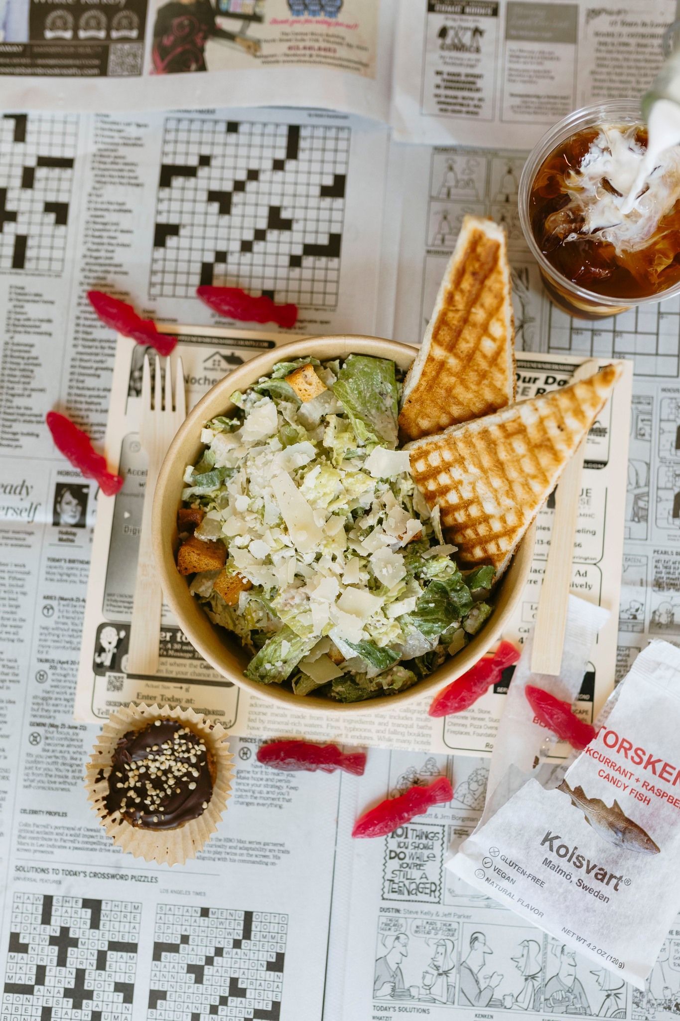 Caesar salad with toast and desserts, surrounded by red gummy fish on newspaper.