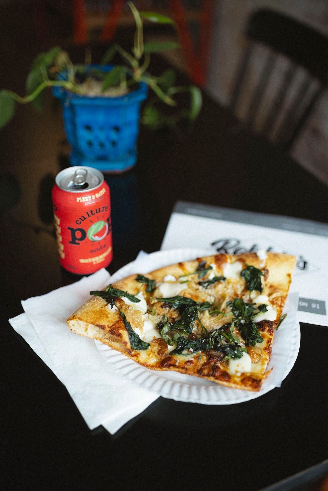 Slice of pizza with spinach, soda can, and plant on a black table.