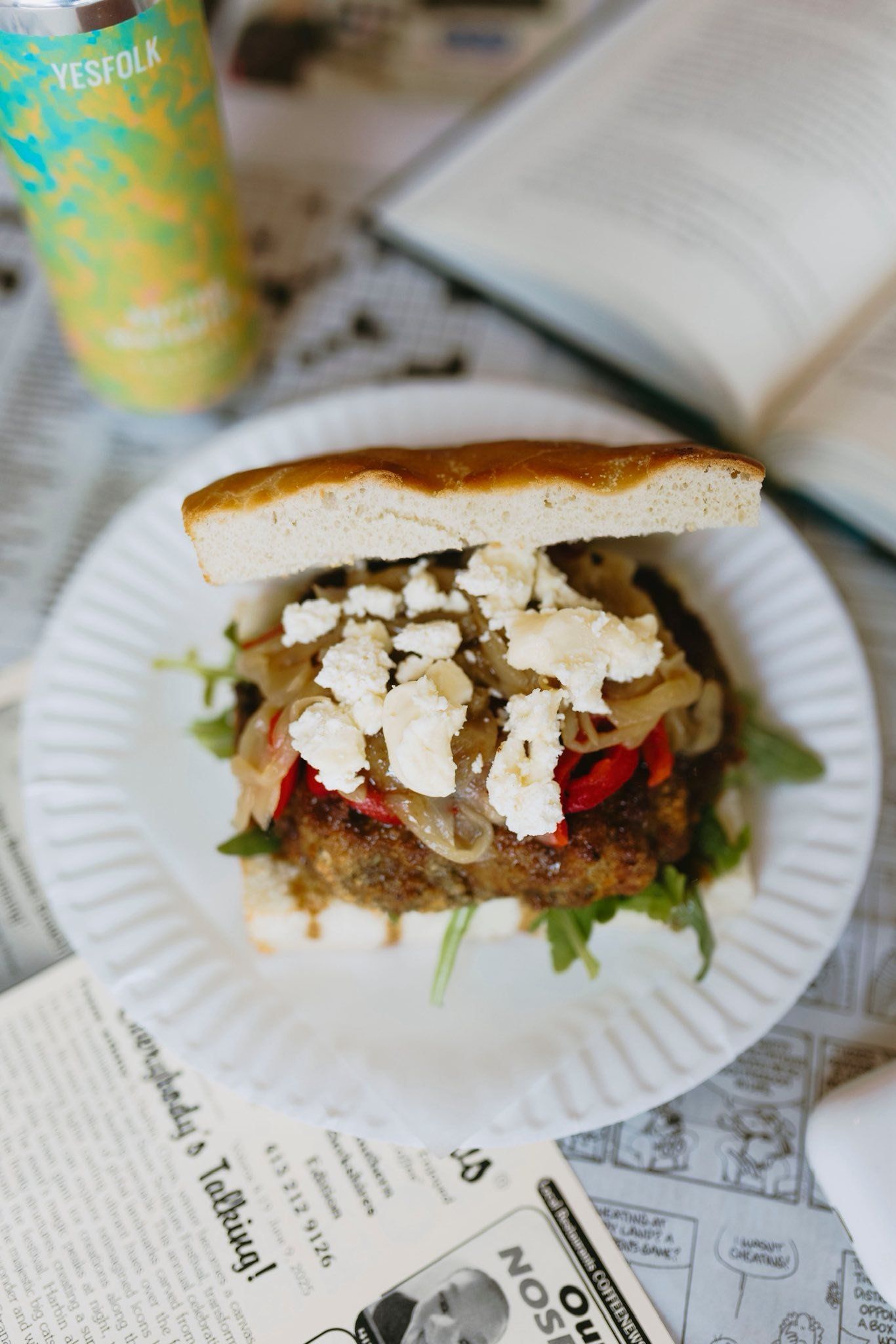 Sandwich on a paper plate, topped with feta and onions, beside a drink and open book on newsprint.