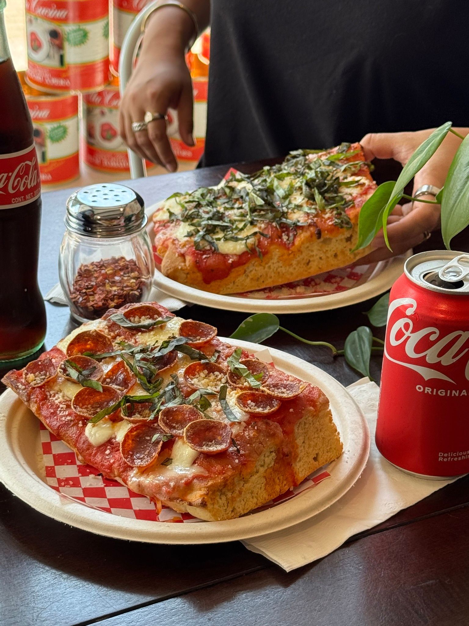Two slices of square pizza on paper plates, with a Coca-Cola and seasonings.