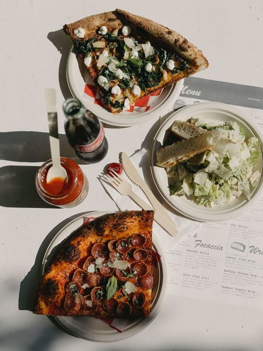 Pizza slices, salad, and a drink on a white table. Includes pepperoni and spinach pizza, red sauce, and wooden utensils.