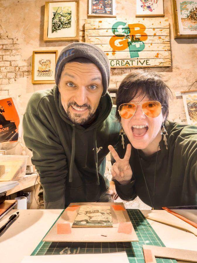 Two smiling people in hoodies pose in a cozy art studio with framed prints and a green cutting mat.