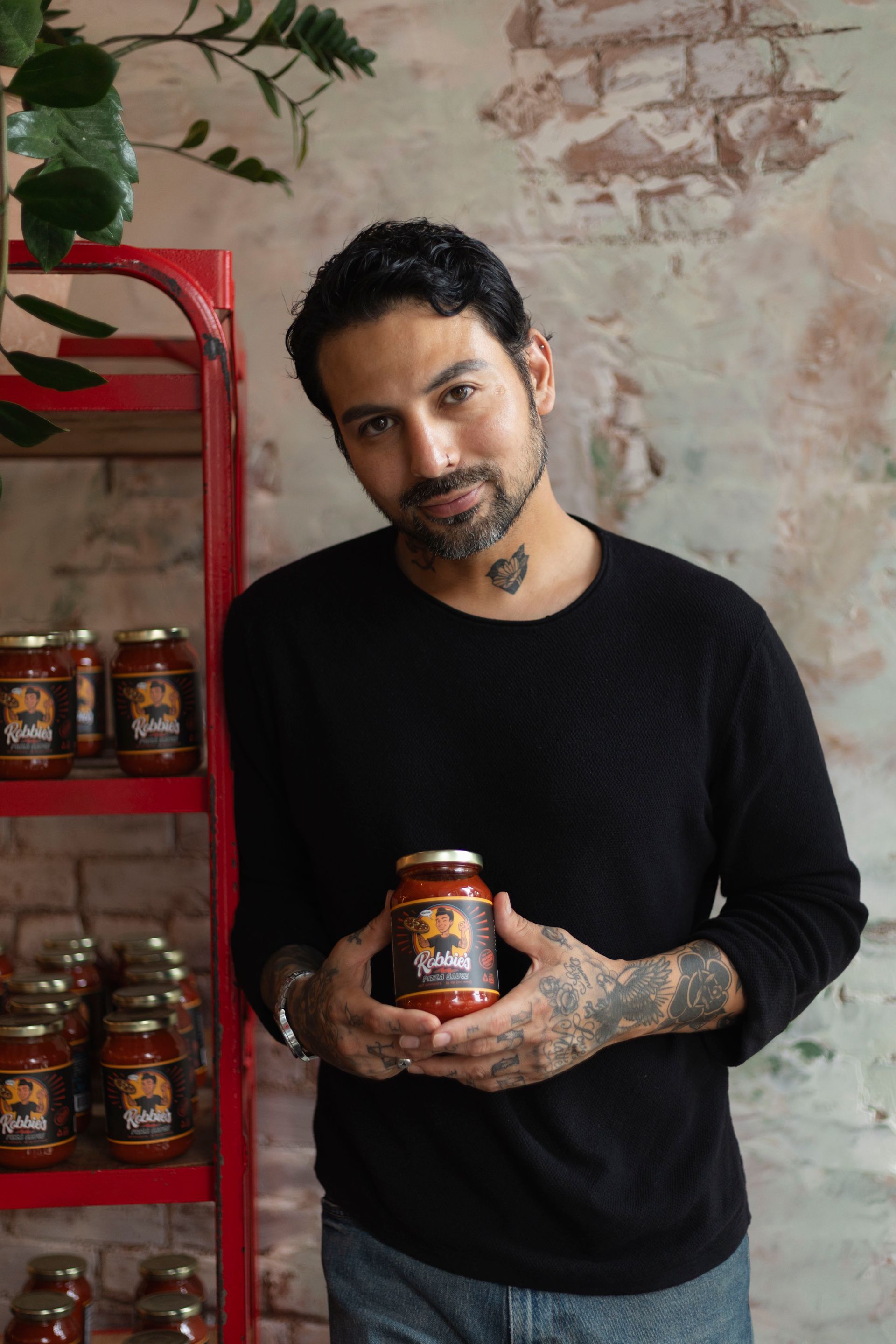 Man with tattoos holding jar of sauce, standing by shelf with jars. Brick wall background.