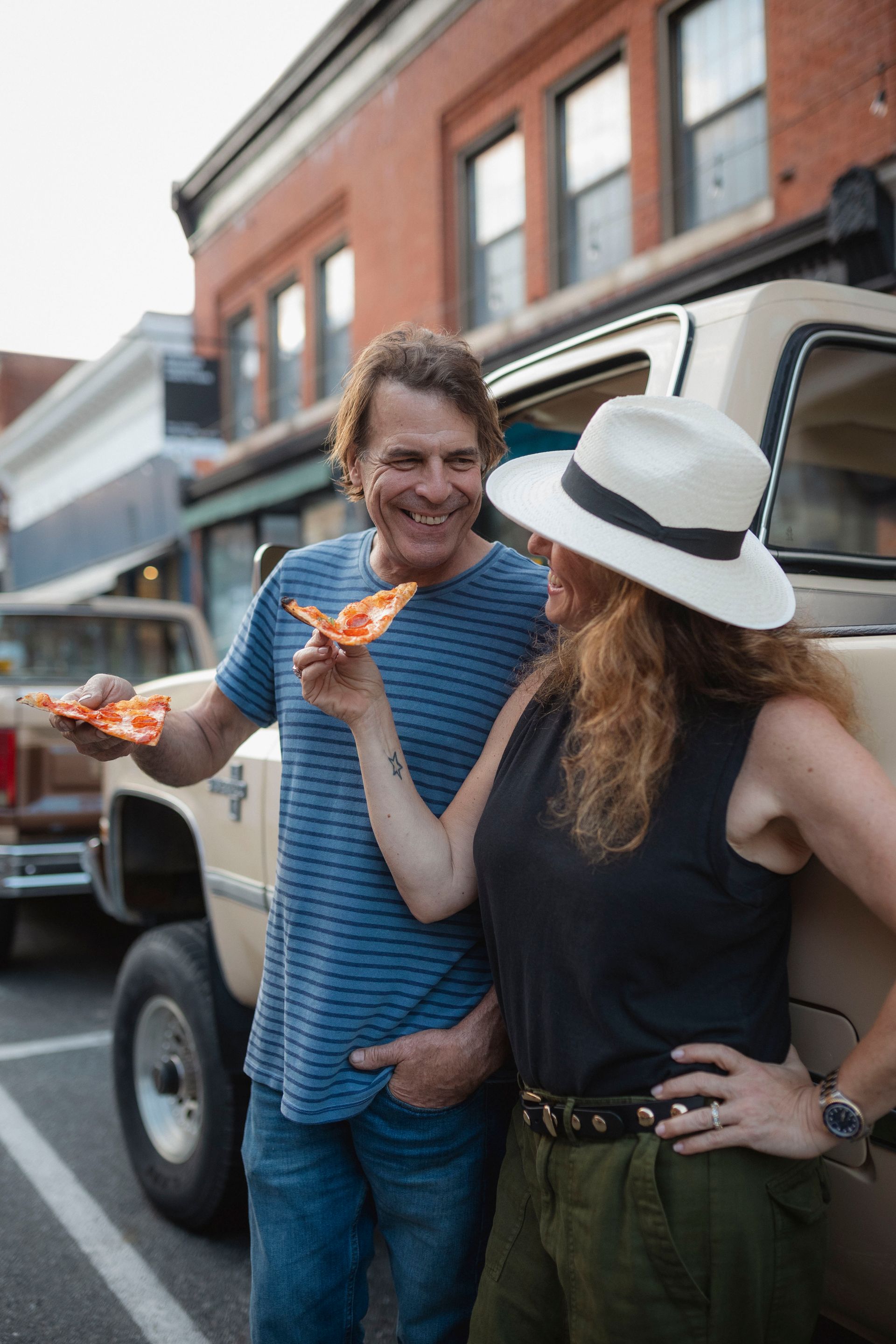 Couple eating pizza near a tan pickup truck, in a small town.