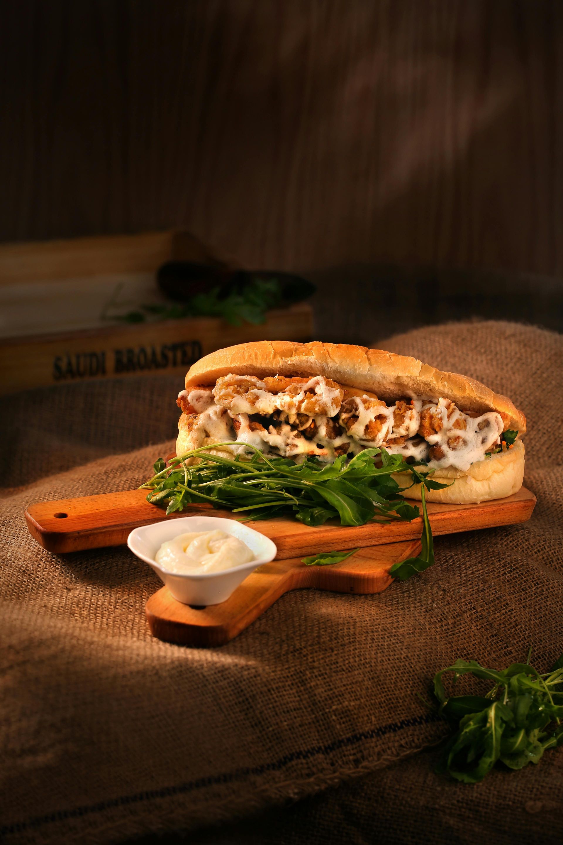 Sandwich on a wooden board with greens, sauce, and sesame seed bun, set on burlap.
