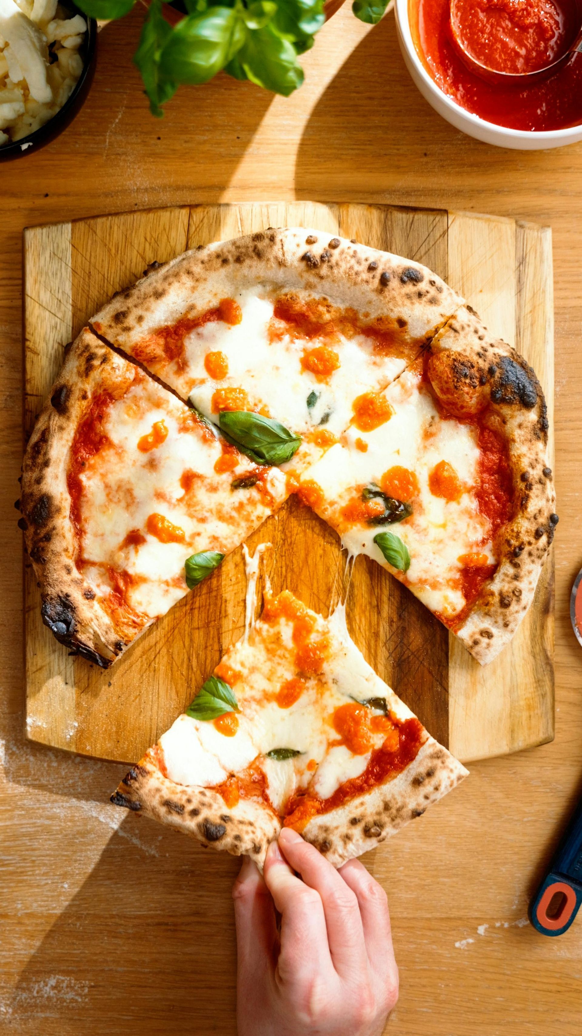 Pizza on a wooden board with a slice being picked up; red sauce, basil, and cheese visible.