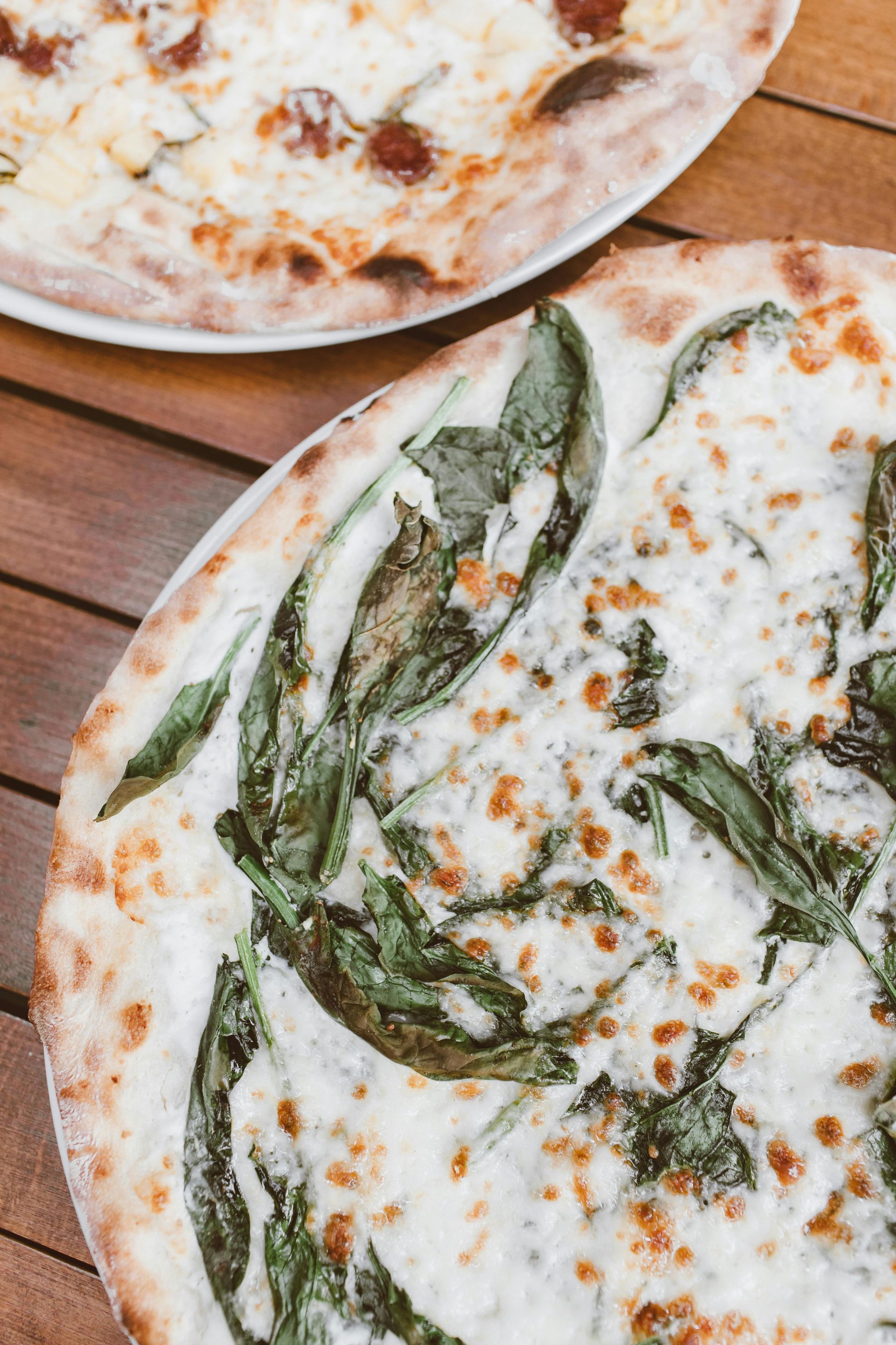 Two pizzas on a wooden table; one with spinach and white sauce, the other with toppings.