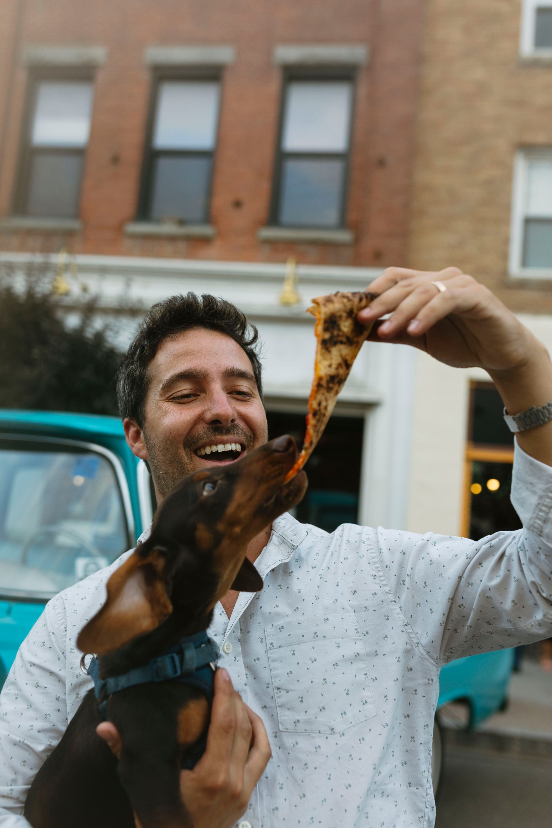 Man and dog sharing pizza slice, smiling. Teal truck and brick building in the background.