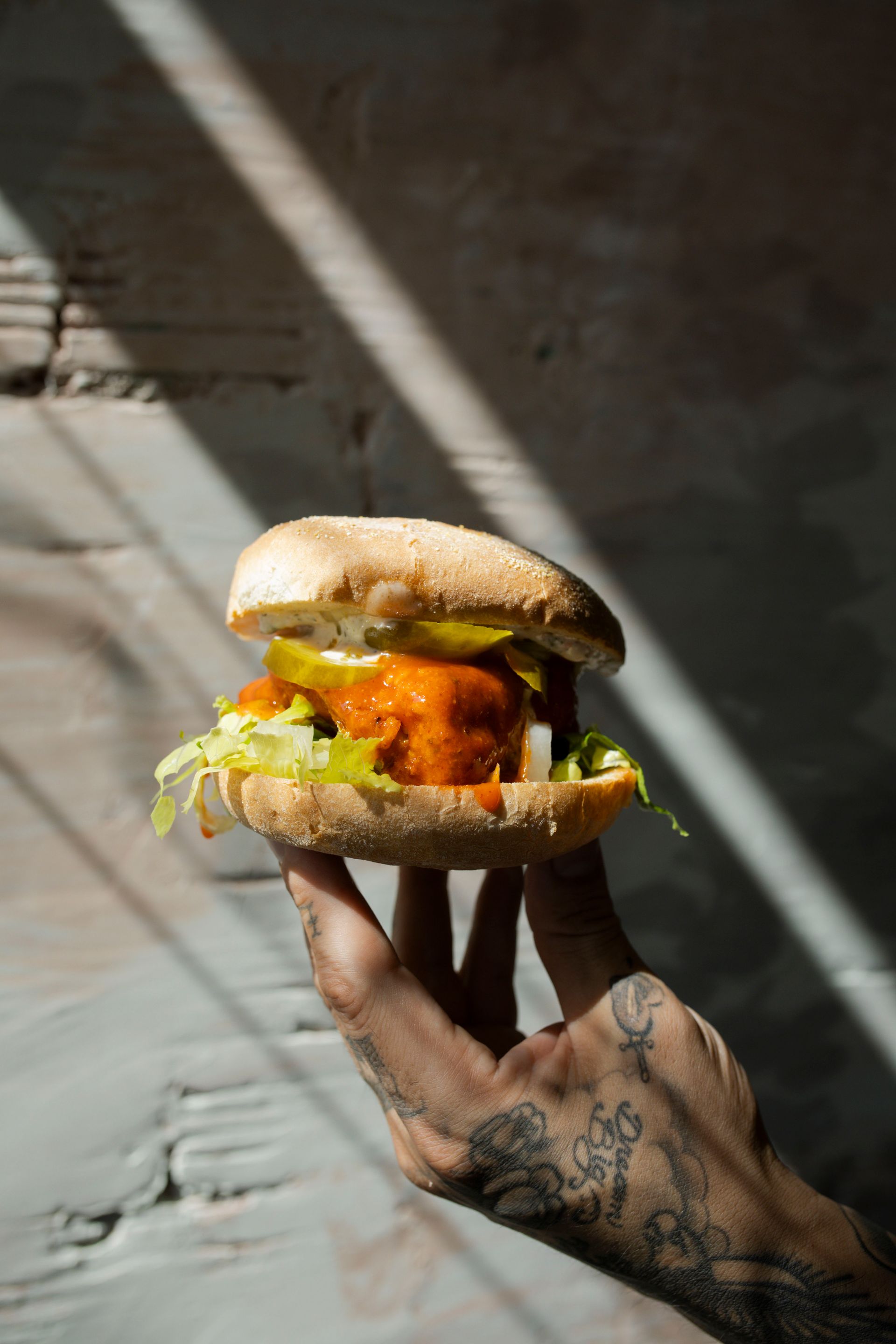 Hand holding a sandwich with a fried patty, lettuce, pickles, and a bun. Tattooed arm against a textured wall.