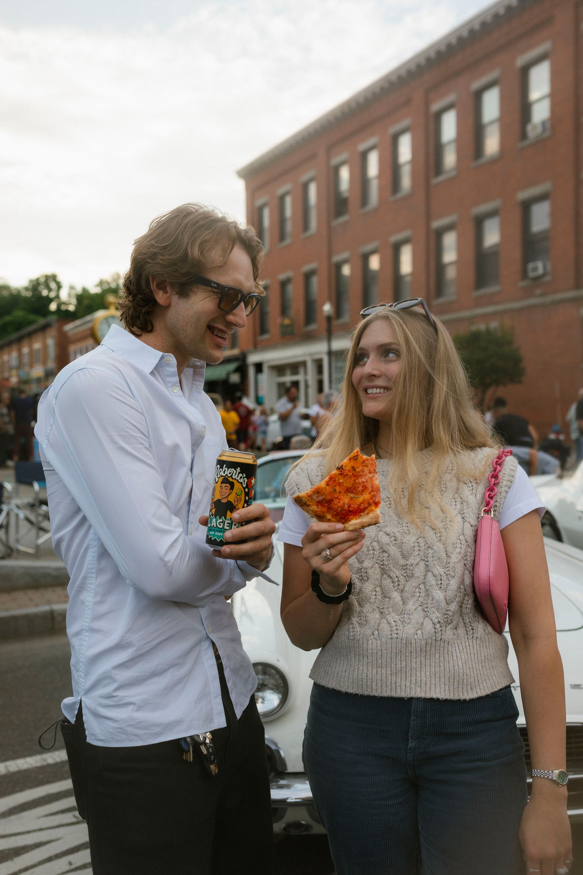 Man and woman smiling, holding pizza and a beverage, standing near a car in a street with brick buildings.