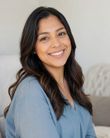 Smiling woman with long dark hair in a light blue top, seated indoors against a soft neutral background