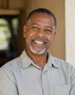 Smiling man in a light gray shirt standing by a doorway, with a blurred background.