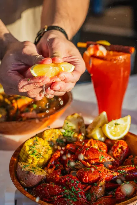 Hands squeezing lemon over a spicy seafood platter beside a red drink on a table