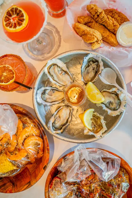 Assorted seafood dishes on a table: oysters, fried fish, shrimp, citrus drinks, and a lobster roll.