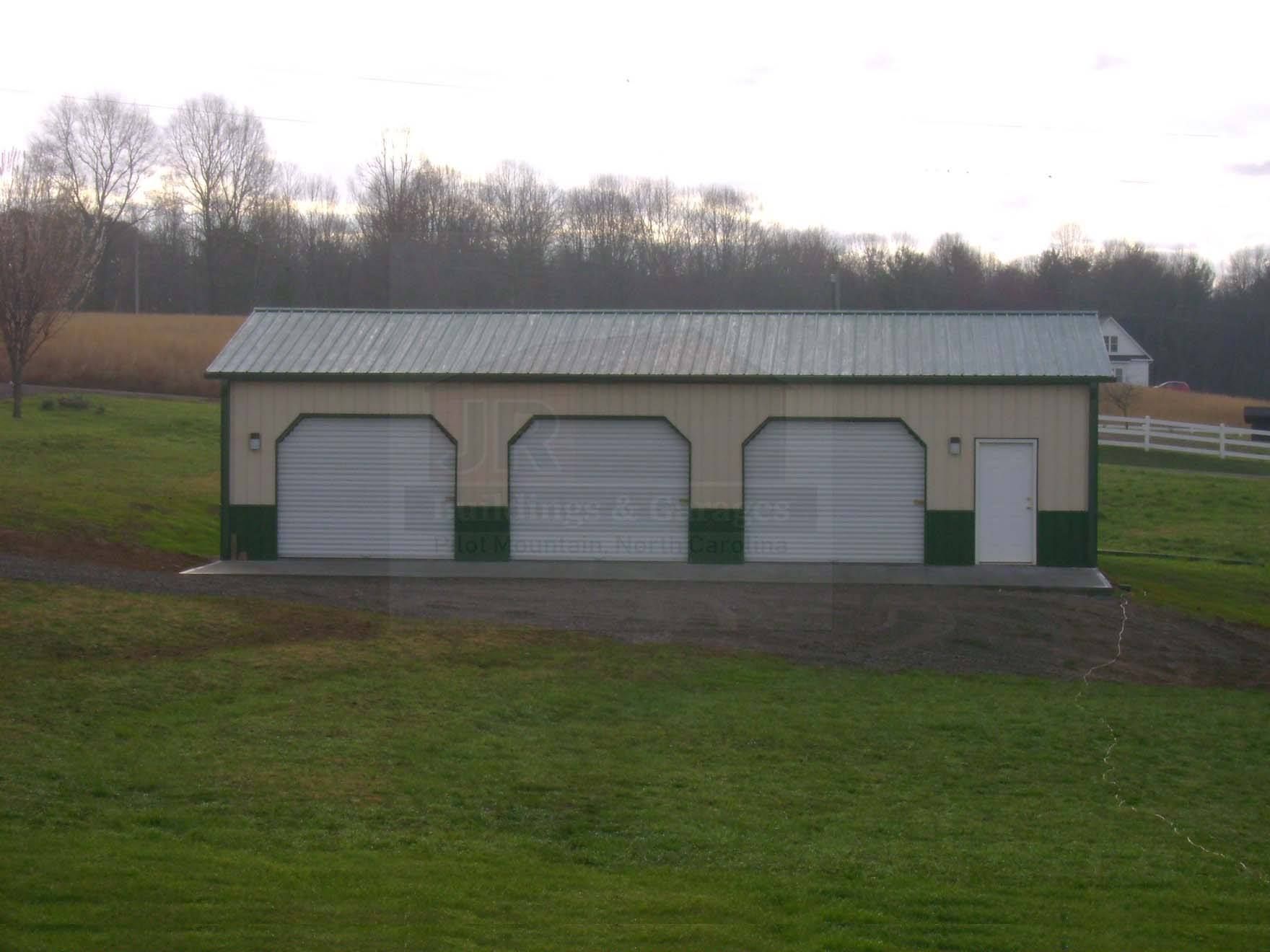 Three-bay garage with green and white accents, situated on a grassy hill, with a treeline in the background.