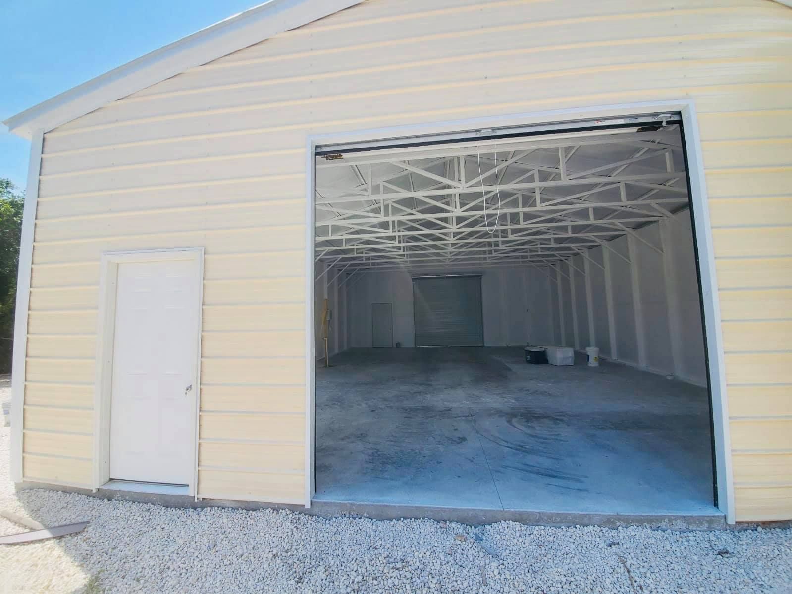 Tan metal building with open garage door, white door to left, gravel in foreground.