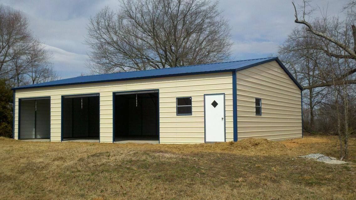 Tan metal building with blue roof, two open garage bays, small door, two windows, set on grassy area.