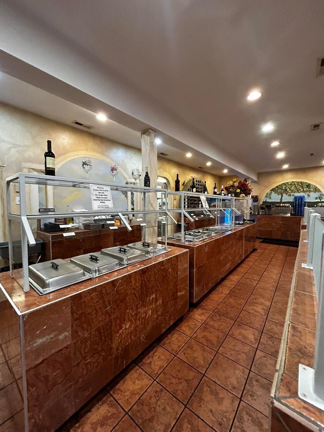 Buffet serving area with stainless steel food containers and shelves. Brown tile floor, wood paneling, and overhead lighting.