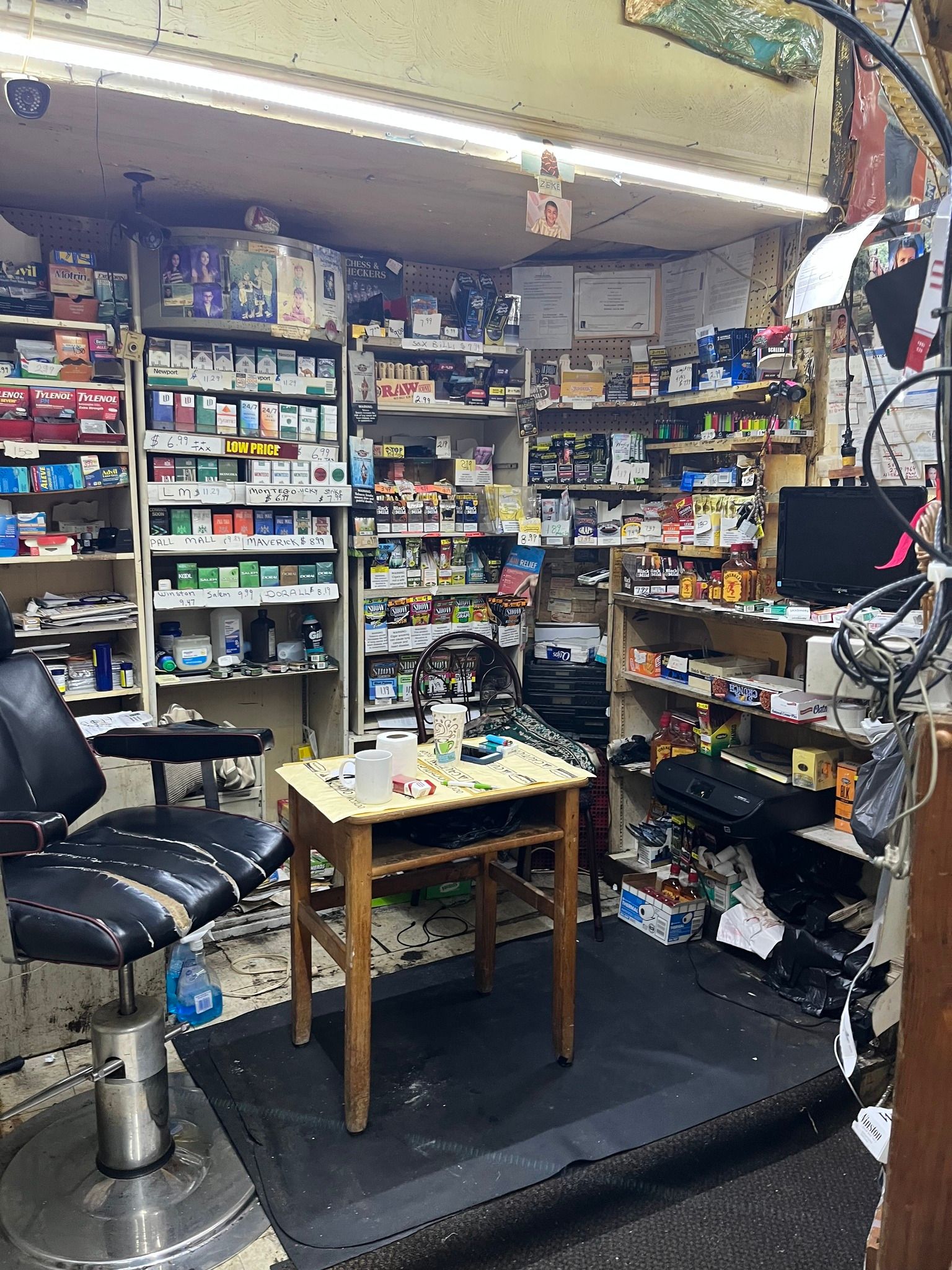 Interior of a cluttered shop, with shelves packed with goods, a small table, a barber chair, and a computer.