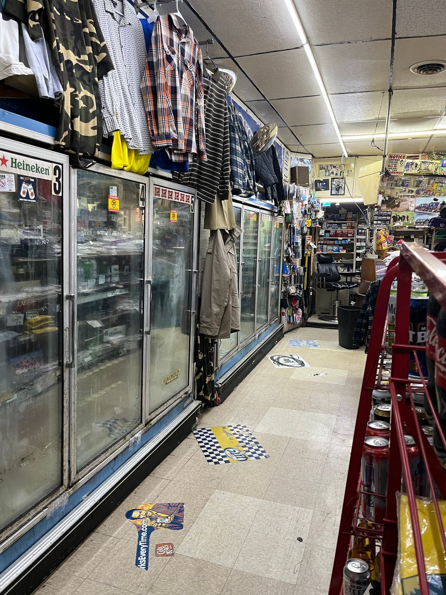 Interior of a convenience store with refrigerated cases, clothing hanging above, and a narrow aisle.