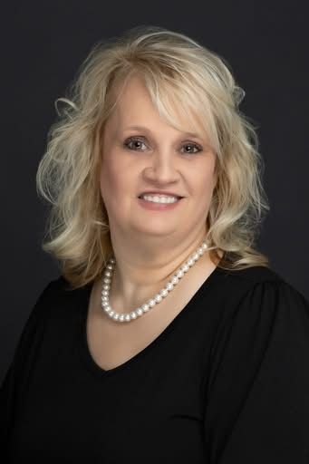 A person with shoulder-length blonde hair wearing a white pearl necklace and a black V-neck top against a black background.