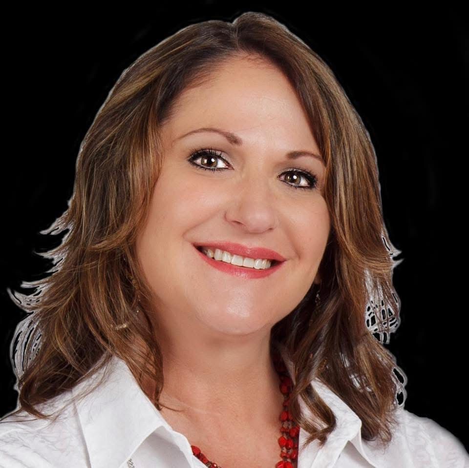 A smiling person with shoulder-length brown hair, wearing a white shirt and red beaded necklace against a black background.