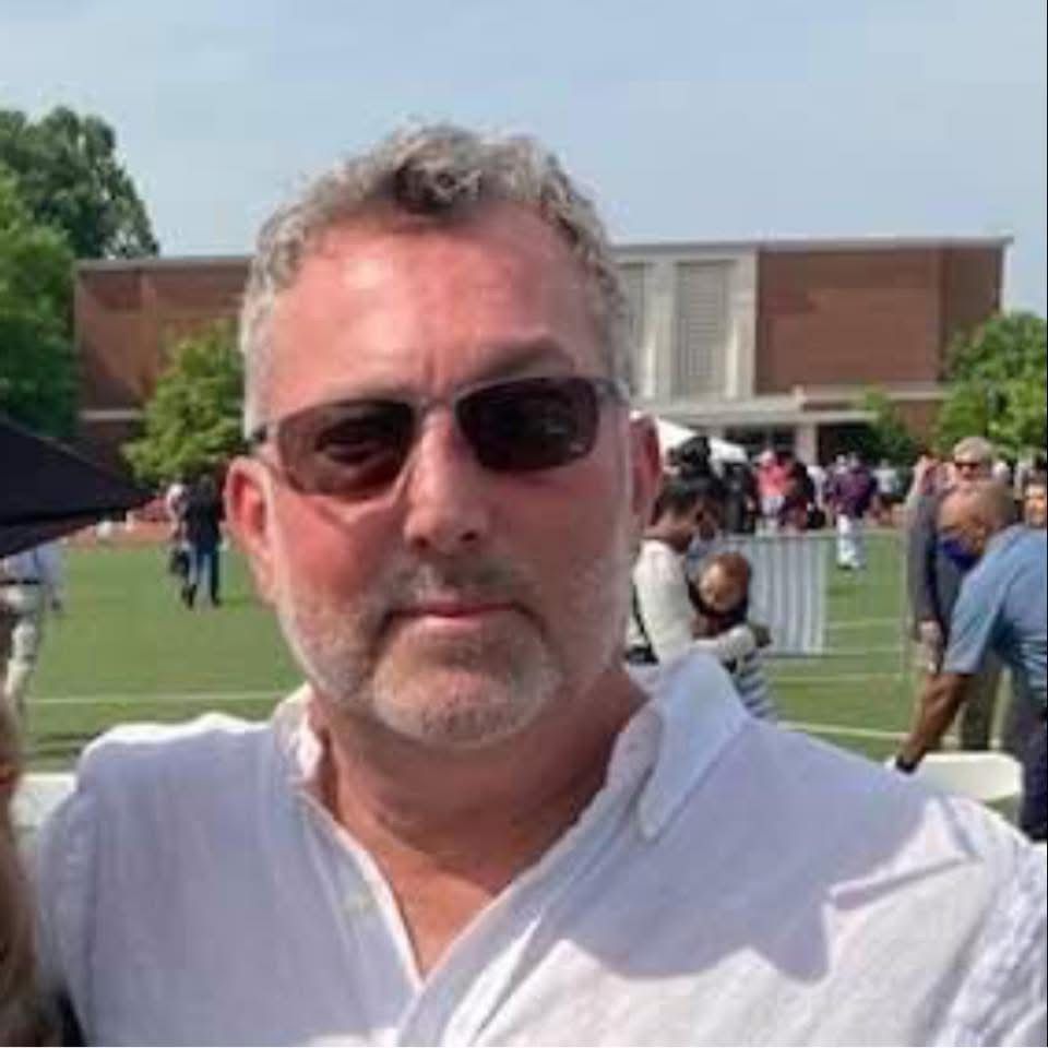A man with grey hair and stubble wearing sunglasses and a white shirt, posing outdoors on a sunny day.