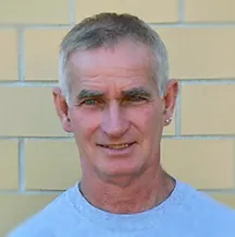 Man with short gray hair and a small earring smiles, standing in front of a brick wall.