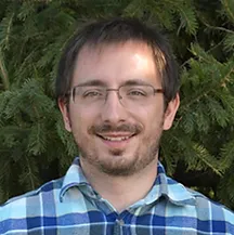 Man with glasses smiling, wearing a blue plaid shirt, set against a green tree backdrop.